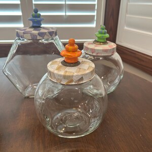 May include: Three clear glass jars with lids. The left jar is hexagonal with a blue and white checkered lid and blue finial. The front jar is round with a tan checkered lid and orange finial. The back jar is round with a pink checkered lid and green finial.