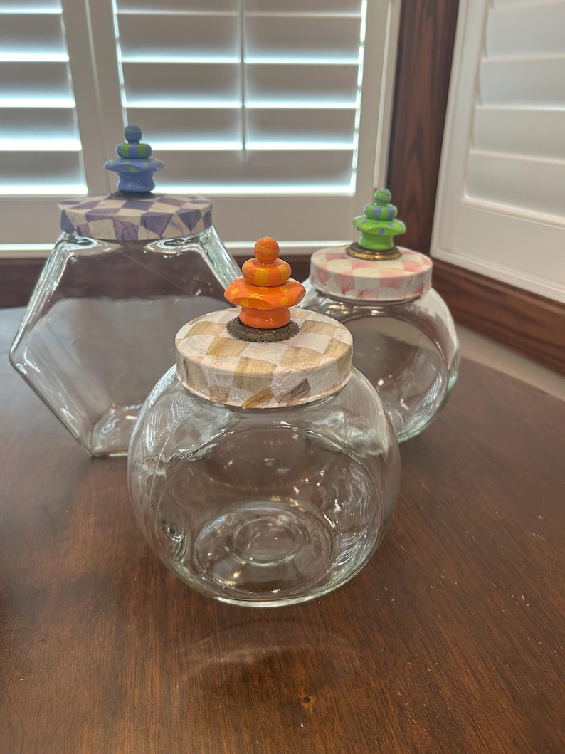 May include: Three clear glass jars with lids. The left jar is hexagonal with a blue and white checkered lid and blue finial. The front jar is round with a tan checkered lid and orange finial. The back jar is round with a pink checkered lid and green finial.