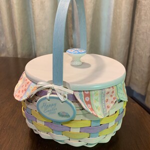 May include: A pastel-colored Easter basket with a light blue handle and lid. The basket is woven with yellow, lavender, and white strips. A tag reads "Happy Easter 2003". The lid has a decorative knob.