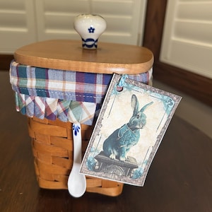 May include: A woven basket with a wooden lid and a fabric trim in blue, green, and red plaid. It has a white ceramic spoon with blue floral accents and a tag with a blue rabbit illustration. The lid has a white ceramic knob with a blue floral design.