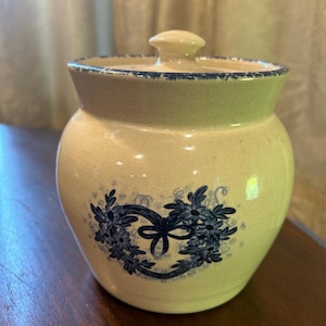 May include: A cream-colored ceramic jar with a blue floral design and a lid. The jar has a blue ribbon and a blue floral wreath design.