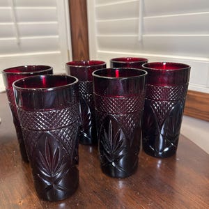 May include: Set of six ruby red drinking glasses. Each glass has a tall, cylindrical shape with a detailed, cut-glass pattern. The glasses are arranged on a wooden surface, showcasing their rich color and design. The glasses are suitable for water, juice, or other beverages.
