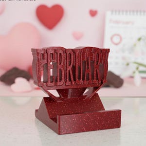 May include: A red speckled stand with the word "FEBRUARY" cut out. The stand is in front of a pink background with heart decorations and a calendar. The stand is designed to hold a phone or other small item.
