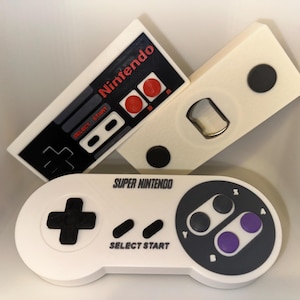 Peut inclure: Trois ouvre-bouteilles blancs et un objet en forme de manette. L'un est un design de manette Nintendo avec des boutons rouges et le mot "Nintendo". Un autre est un design de manette Super Nintendo avec les mots "Super Nintendo" et "Select Start".