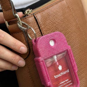 Op de afbeelding: Een felroze glitterende siliconen hoesje met een rechthoekige Touchland Power Mist hydraterende handdesinfectiespray. Het hoesje is voorzien van een metalen ring die is bevestigd aan de riem van een bruine leren handtas met textuur.
