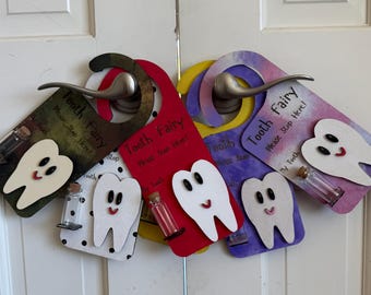 May include: A collection of five colorful door hangers, each featuring a white tooth cutout with a smiling face, the text Tooth Fairy Please Stop Here, and a small glass vial with a cork stopper attached for holding a lost tooth.