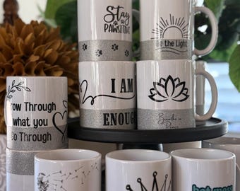 May include: White ceramic mugs with various black and teal designs and text. Designs include a paw print, a sun, a lotus flower, and the phrases "Stay PAWSITIVE", "Be the Light", "I AM ENOUGH", and "hot mess but caffeinated".