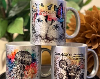 May include: Three silver mugs with unique designs. One mug features two fluffy kittens with butterflies. Another has a colorful jellyfish design. The third mug displays a Highland cow surrounded by sunflowers and the text "Deja-Moo".