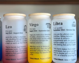 May include: Three glass jars with wooden lids, each labeled with a different zodiac sign: Leo, Virgo, and Libra. The jars have a frosted appearance and contain text describing each sign's characteristics. The jars are on a wooden shelf.