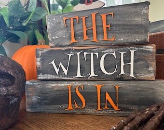 May include: A stack of three rectangular wooden blocks with a distressed dark gray finish. The blocks feature raised lettering that reads THE WITCH IS IN in orange and white. A wooden witch hat cutout sits atop the stack with a black band.