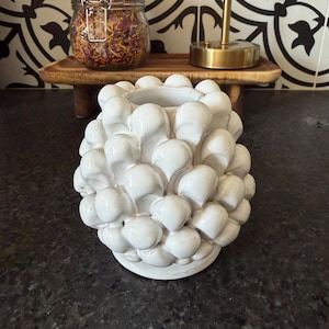 May include: A white ceramic vase with a textured, bubble-like design. The vase is round and sits on a dark countertop. In the background, a wooden tray holds a jar with a metal clasp and a gold-colored lamp.