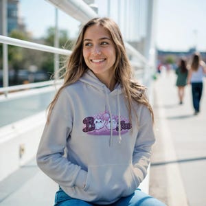 May include: A light gray hooded sweatshirt featuring a graphic print on the chest. The design displays two small white ghosts with pink bows, a disco ball, and the text "Boo" in a dark purple, glittery, plaid-patterned font with sparkles.