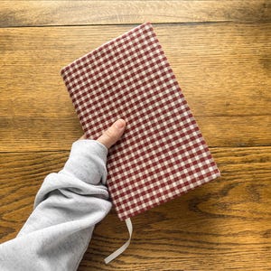 May include: A rectangular notebook featuring a fabric cover with a red and white gingham check pattern. The notebook includes a thin white ribbon bookmark extending from the bottom edge, showcasing a classic textile design and structured form.