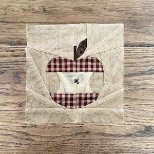 May include: A square fabric quilt block featuring a pieced apple design. The apple is constructed from red and white gingham fabric with a cream-colored center and a dark brown stem. The surrounding background consists of textured beige fabric.