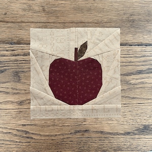May include: A square fabric quilt block featuring a dark maroon apple with a small brown stem and a single dark brown leaf, centered against a light beige background with a subtle tonal pattern, constructed using pieced fabric segments.