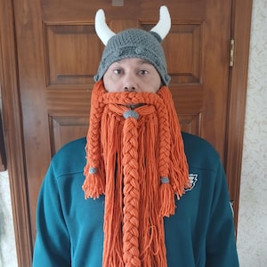 May include: A gray knitted Viking hat with white horns and a long orange braided beard.