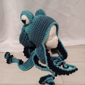 May include: A teal and dark navy blue crocheted hat featuring two large, round eyes on top and multiple long, dangling tentacles. The hat is constructed with a textured yarn pattern, incorporating dark navy blue accents along the edges of the tentacles.
