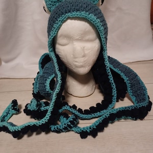 May include: A crochet hat featuring a dark blue base with a light blue trim and two protruding frog-like eyes on top. The hat includes long, dangling tentacles in dark blue and light blue, each adorned with small, rounded, dark blue bobble accents.