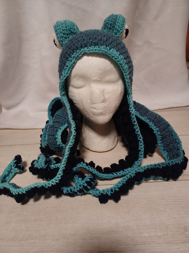 May include: A crochet hat featuring a dark blue base with a light blue trim and two protruding frog-like eyes on top. The hat includes long, dangling tentacles in dark blue and light blue, each adorned with small, rounded, dark blue bobble accents.