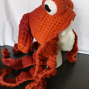 May include: A handmade crochet hat in a vibrant orange hue, designed to resemble an octopus. The hat features two large, round eyes with white sclera and black pupils, and several long, textured, dangling tentacles that extend from the base.