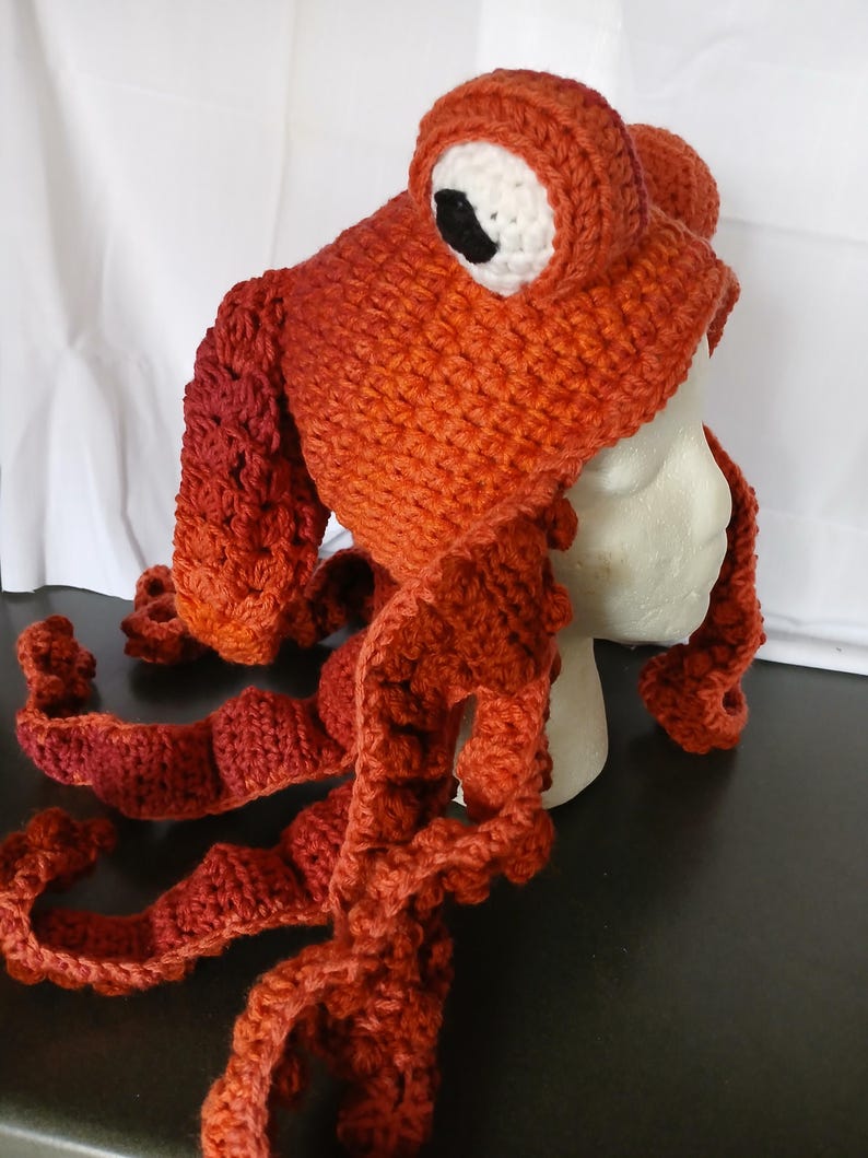 May include: A handmade crochet hat in a vibrant orange hue, designed to resemble an octopus. The hat features two large, round eyes with white sclera and black pupils, and several long, textured, dangling tentacles that extend from the base.