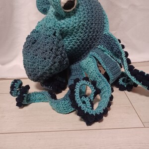 May include: A handmade crochet octopus hat in shades of teal and dark blue. The hat features a large, singular white and black eye on the front, with multiple long, curling tentacles extending downwards, each accented with dark blue bobble stitches.