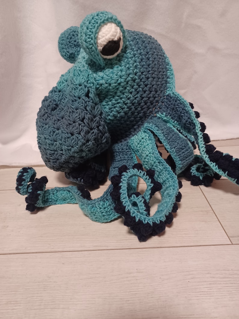 May include: A handmade crochet octopus hat in shades of teal and dark blue. The hat features a large, singular white and black eye on the front, with multiple long, curling tentacles extending downwards, each accented with dark blue bobble stitches.