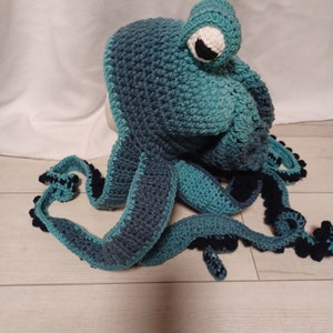May include: A crochet hat shaped like an octopus in shades of teal and dark blue. The hat features a large, protruding eye with a white sclera and a black pupil, and several long, dangling tentacles with dark blue undersides and textured edges.