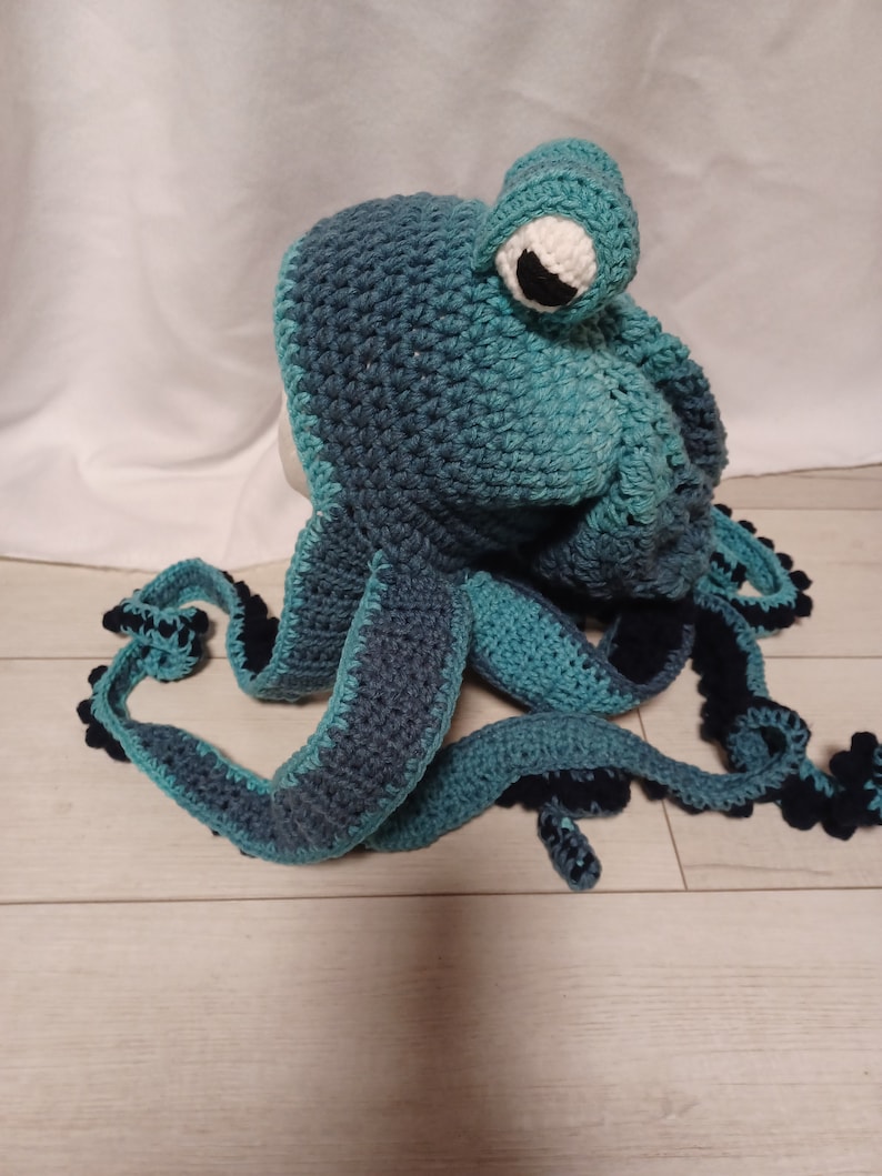 May include: A crochet hat shaped like an octopus in shades of teal and dark blue. The hat features a large, protruding eye with a white sclera and a black pupil, and several long, dangling tentacles with dark blue undersides and textured edges.