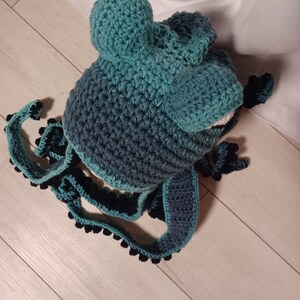 May include: A teal and dark blue crocheted hat featuring two rounded protrusions on top and long, dangling side panels. The edges of the side panels are decorated with small, dark blue crocheted spheres, creating a textured, octopus-like appearance.