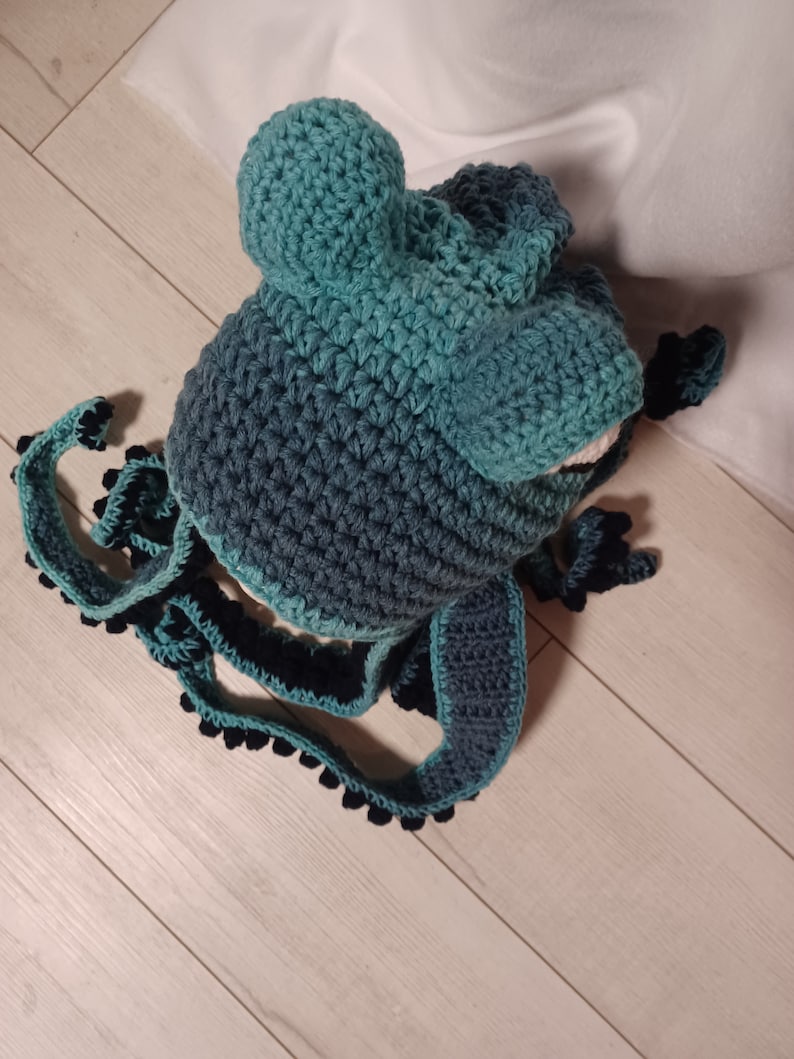 May include: A teal and dark blue crocheted hat featuring two rounded protrusions on top and long, dangling side panels. The edges of the side panels are decorated with small, dark blue crocheted spheres, creating a textured, octopus-like appearance.