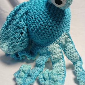 May include: A handmade crochet octopus toy crafted from bright blue yarn. The creature features a large, rounded head, a single prominent eye with a white border and black pupil, and multiple curling tentacles in alternating shades of blue.