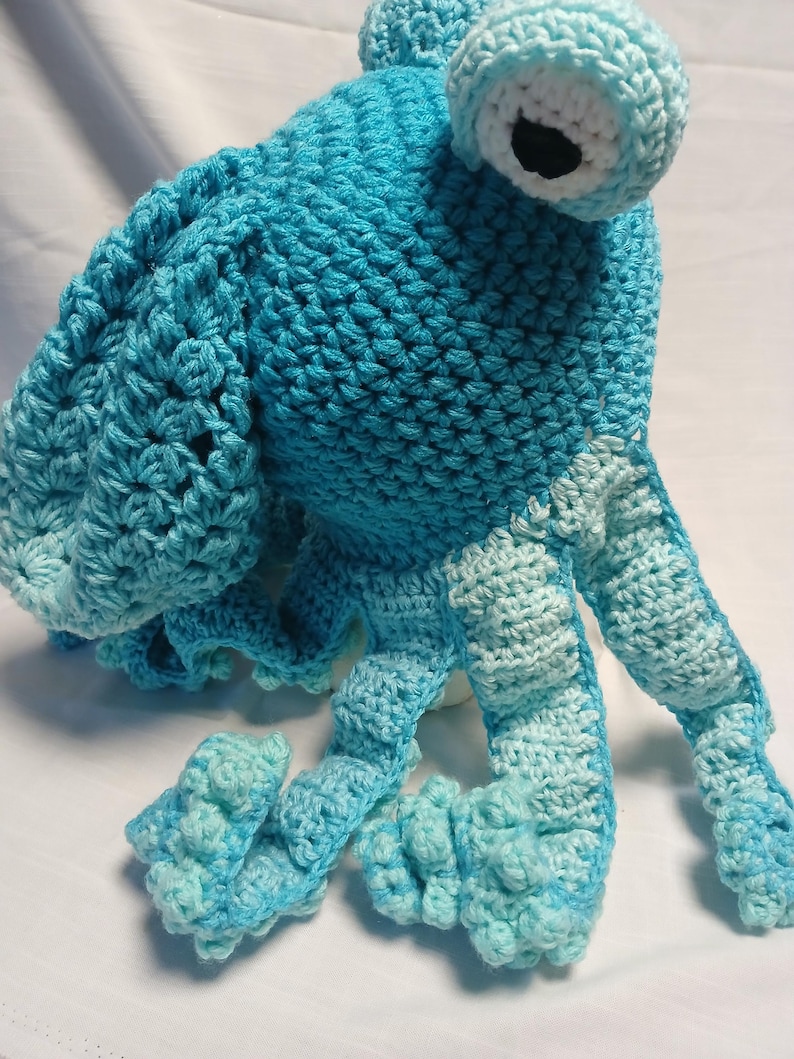 May include: A handmade crochet octopus toy crafted from bright blue yarn. The creature features a large, rounded head, a single prominent eye with a white border and black pupil, and multiple curling tentacles in alternating shades of blue.