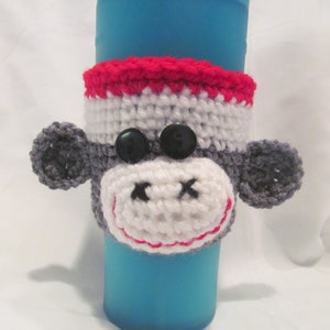 Puede incluir: Un calentador de taza de café de crochet con forma de cara de mono con hilo rojo, blanco y gris. El calentador tiene dos botones negros para los ojos.