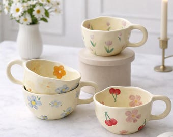 Puede incluir: Cuatro tazas de cerámica color crema con diseños florales. Una taza presenta cerezas, otra flores naranjas y las otras flores azules y rosas. Cada taza tiene un asa y un borde ondulado. Un jarrón de flores blancas y una vela encendida están en el fondo.