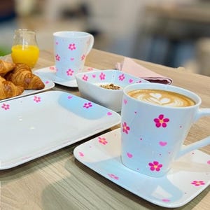 Peut inclure: Service de table en céramique blanche orné de fleurs et de cœurs roses. Comprend des assiettes, des bols et des tasses avec soucoupes. Un verre de jus d'orange et des croissants sont également sur la table.