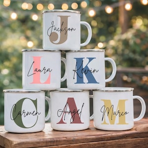 Peut inclure: Mugs en émail blanc avec des bords argentés, personnalisés avec un nom et une initiale de différentes couleurs. Les noms incluent "Jackson", "Laura", "Kevin", "Carmen", "Angel" et "Marco". Les tasses sont empilées.