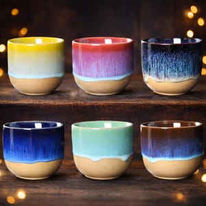Op de afbeelding: Zes keramische bekers met een uniek ontwerp. Elke beker heeft een kleurenschema met verloop, dat overgaat van levendige tinten naar een neutrale basis. De kleuren zijn geel, paars, blauw, groen en bruin. De bekers zijn op een houten oppervlak geplaatst.