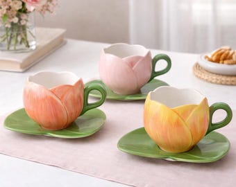 Puede incluir: Tres tazas de té de cerámica con forma de tulipán con platillos a juego. Las tazas son en tonos rosa, naranja y amarillo, con asas y platillos verdes. Un jarrón de flores rosas está en el fondo.