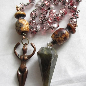 Puede incluir: Un collar largo con un colgante de cristal de punta verde oscuro, una pequeña figura de mujer de bronce y cuentas de cerámica marrón y blanca. El collar está hecho de cuentas de vidrio transparente con motas rojas.