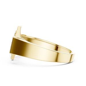May include: A gold-colored ring with a unique design. The ring has a wide band and a detailed top, possibly featuring a stylized shape. The ring is shown on a white background, highlighting its polished surface and intricate details.