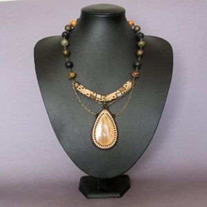 May include: A multi-layered necklace with a teardrop-shaped pendant. The pendant is made of a light brown stone with a beaded border. The necklace is made of brown, black, and green beads, and has a gold chain.