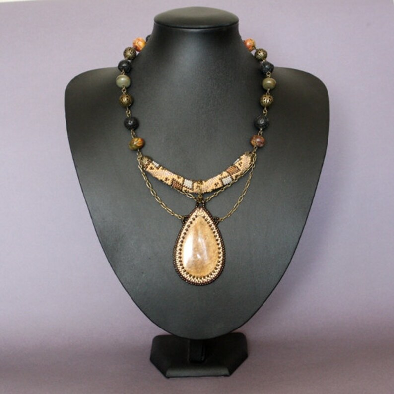 May include: A multi-layered necklace with a teardrop-shaped pendant. The pendant is made of a light brown stone with a beaded border. The necklace is made of brown, black, and green beads, and has a gold chain.