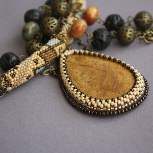May include: A beaded necklace with a brown stone pendant. The necklace features a variety of beads, including brown, black, and white beads. The pendant is surrounded by a beaded border.
