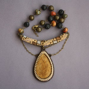May include: A beaded necklace with a teardrop-shaped pendant. The pendant is made of brown leather and is surrounded by a beaded border. The necklace is made of brown, black, and white beads. The necklace is on a gray background.