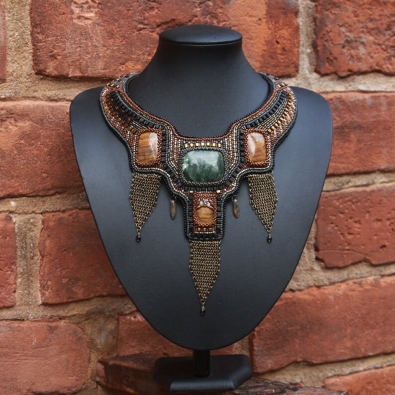 May include: A beaded necklace with a geometric design featuring green, brown, and black stones. The necklace is displayed on a black mannequin bust.