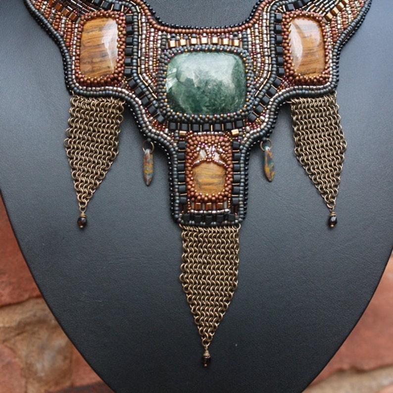 May include: A beaded necklace with a geometric design featuring tiger's eye and green stones. The necklace has three long chains hanging from the bottom.