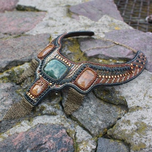 May include: A beaded statement necklace with a green, brown, and black colour scheme. The necklace features a large green stone and smaller brown stones. The necklace is accented with gold chain fringe.