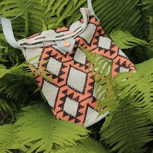 May include: A handmade crochet bag featuring a geometric pattern in peach, brown, and white. It has a light blue strap and a button closure. The bag is set amongst green fern leaves.