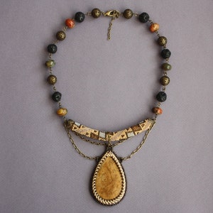 May include: A beaded necklace with a teardrop-shaped pendant. The pendant is made of a light brown material and is surrounded by a beaded border. The necklace is made of brown, black, and green beads.
